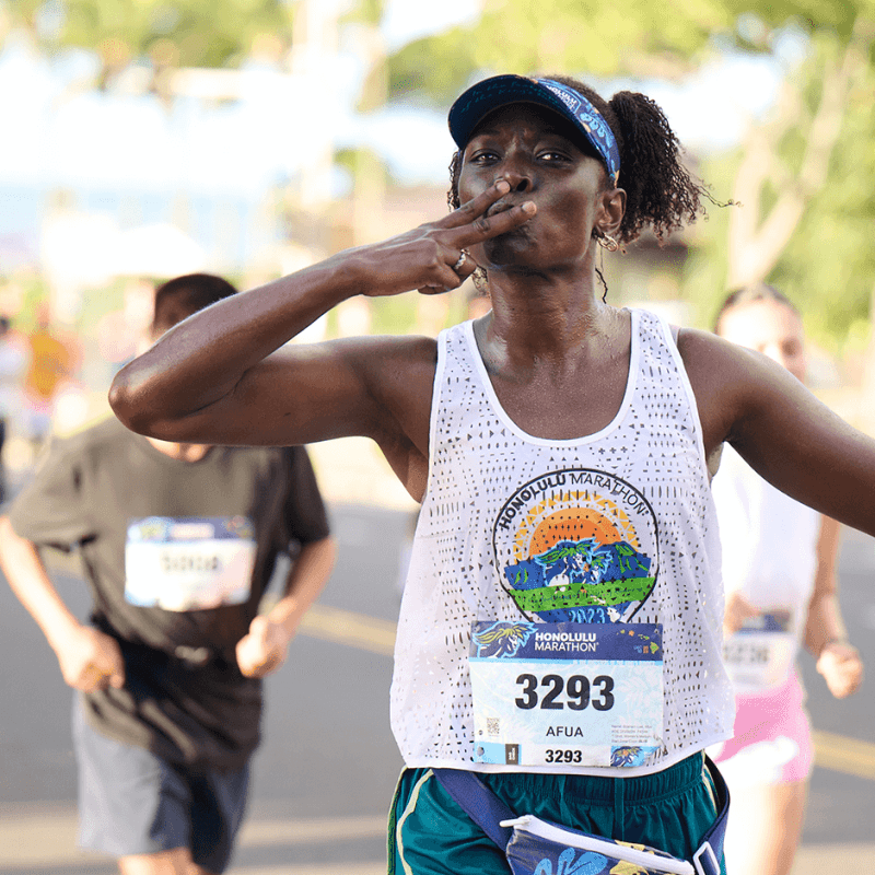 Runner in a marathon blowing a kiss, wearing a white tank top and a cap on a sunny day.