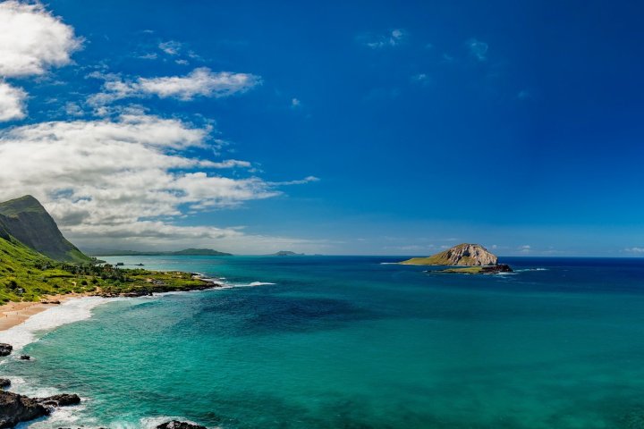 Scenic coastal landscape with blue ocean, green mountains, and an island under a clear blue sky.