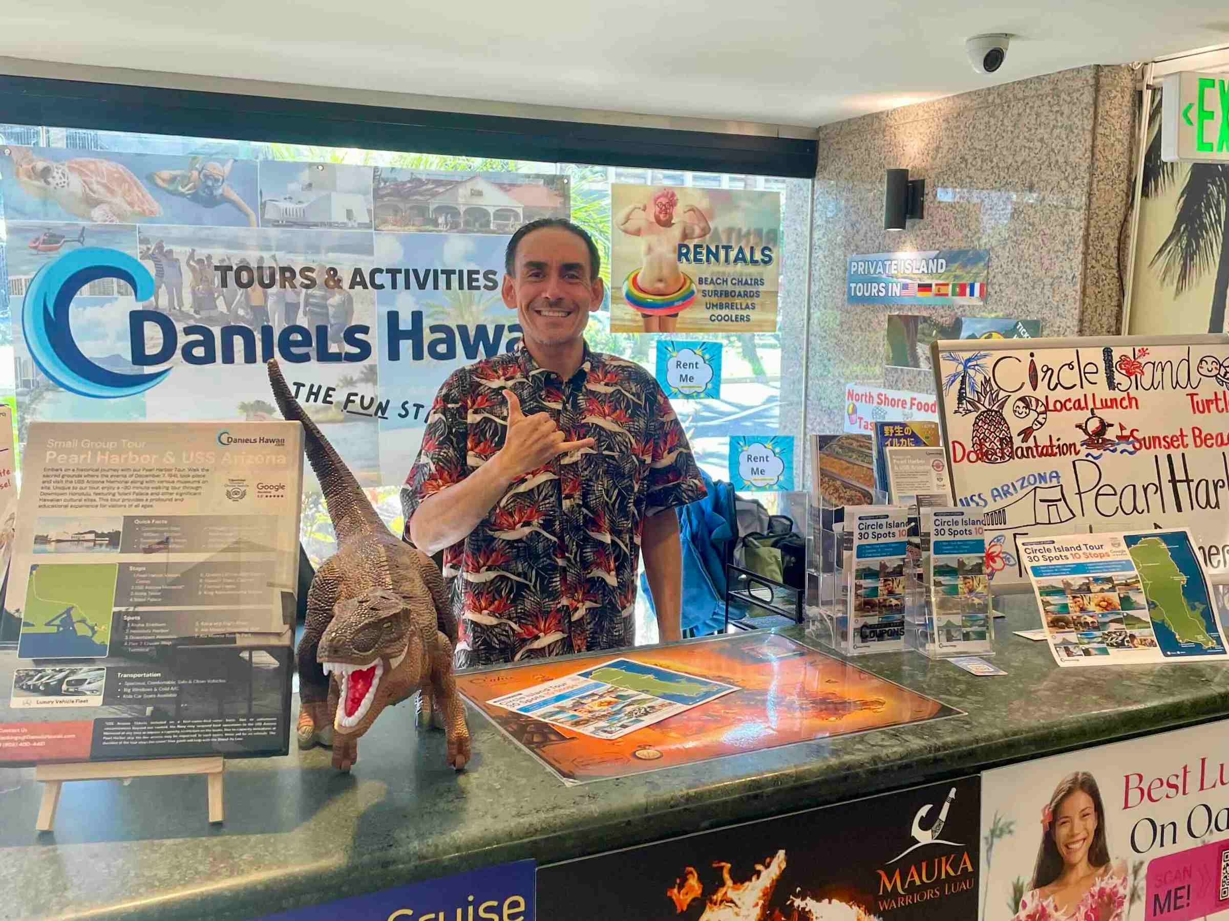 Man in floral shirt behind tourist counter with brochures and dinosaur toy.